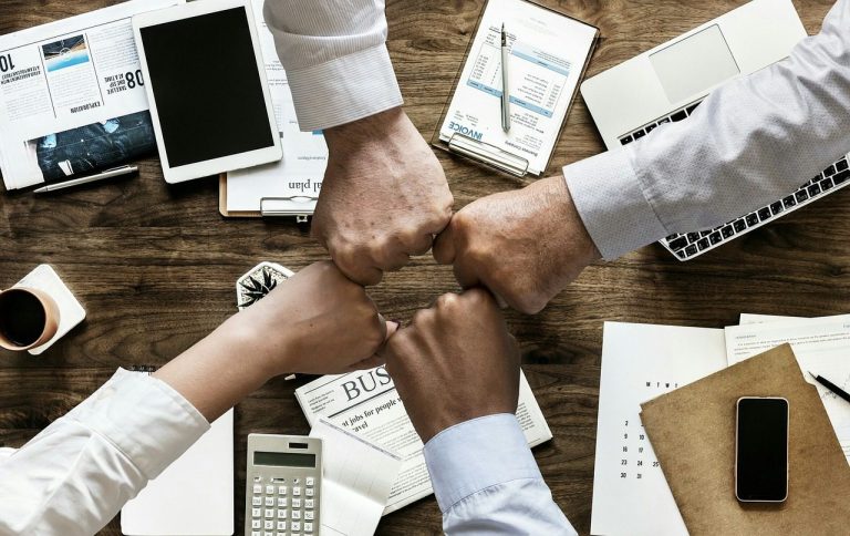 Quatre mains se rencontrent au centre d'une table, entourées de documents et d'appareils.