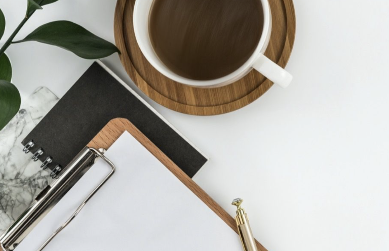 Tasse de café sur une soucoupe en bois, carnet et stylo sur une table blanche.